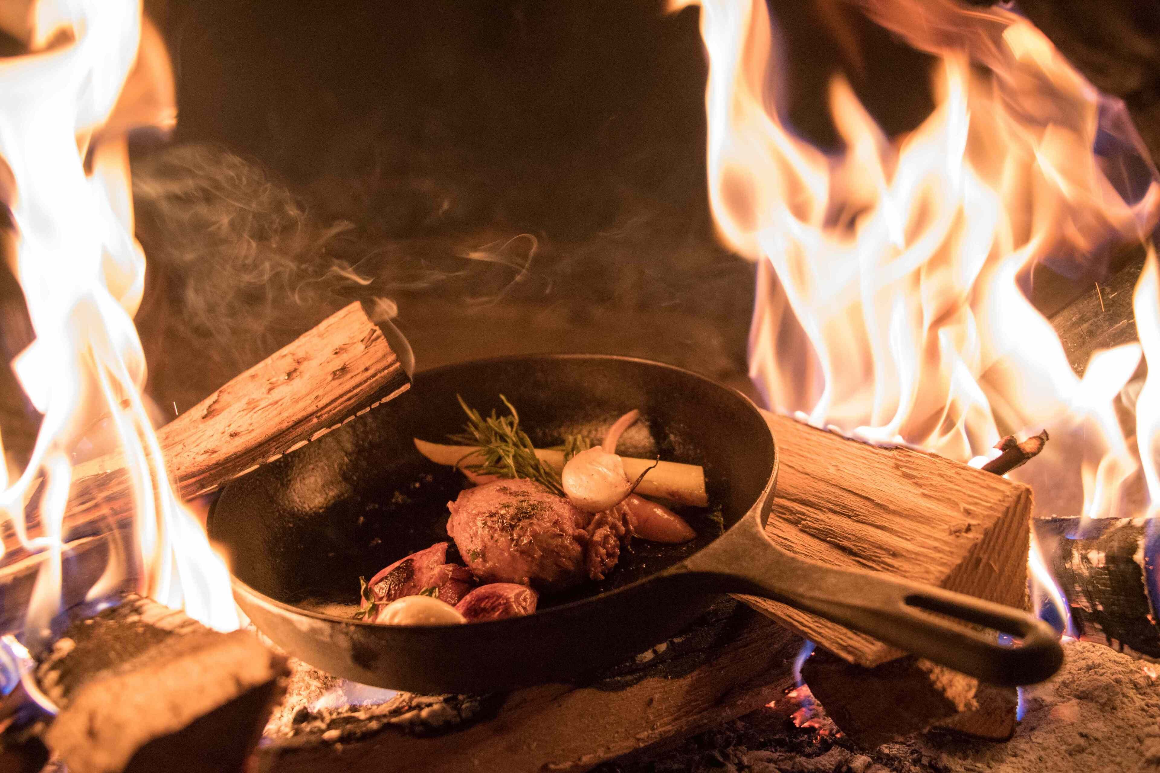 Une poêle contenant des aliments, placée au-dessus d’une flamme vive et entourée de bois en combustion.