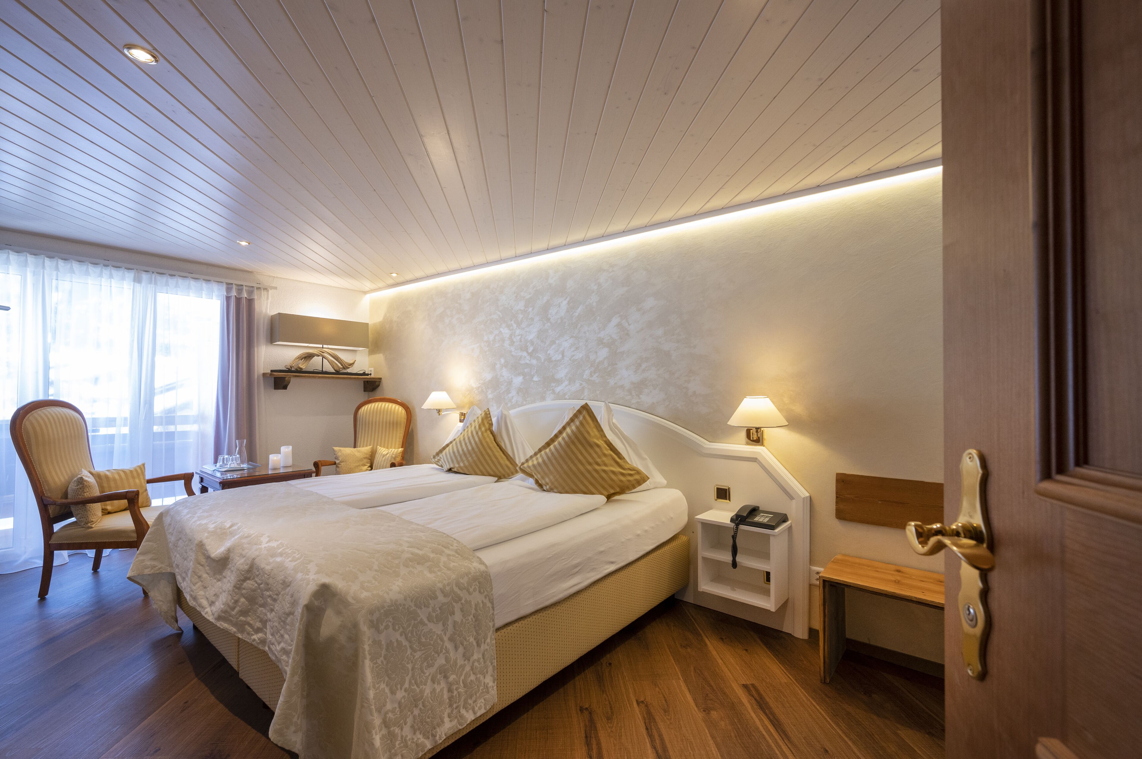 Habitació d'hotel amb un llit gran, dues taules de nit, una cadira, una panxa de fusta i una porta amb manilla de bronze.