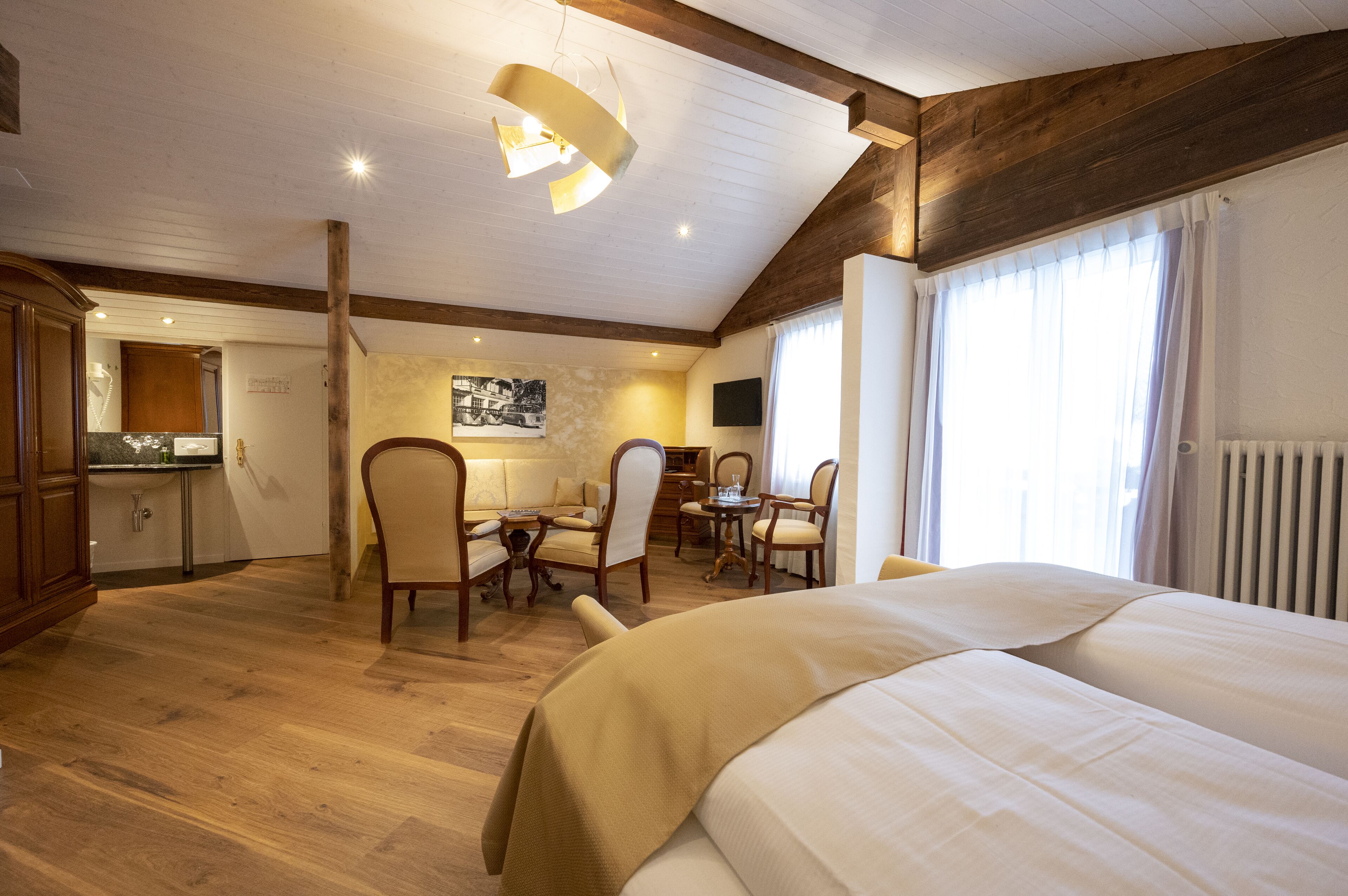 Ein Hotelzimmer mit einem großen Bett, einer Essecke mit Stühlen und Tisch, einer Küchenzeile und einem großen Fenster mit Vorhängen.