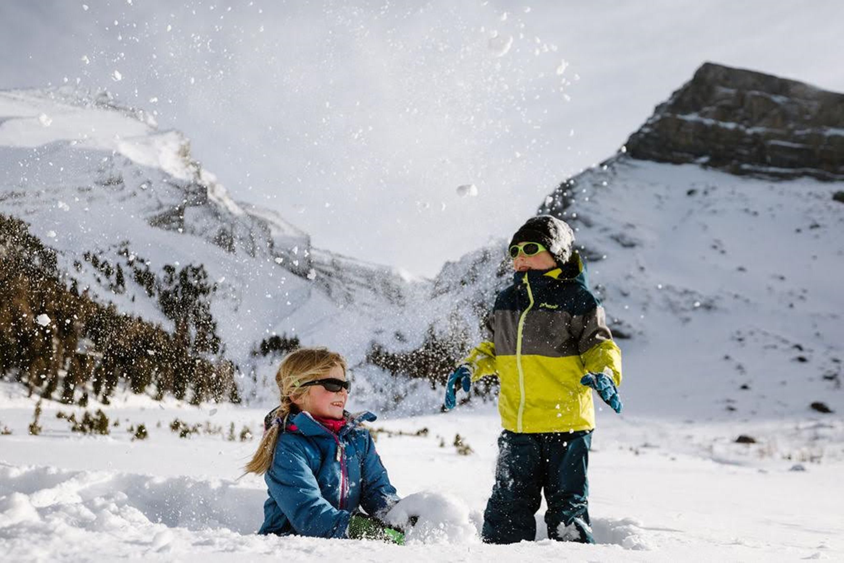 Dos niños jugando en la nieve con montañas al fondo.