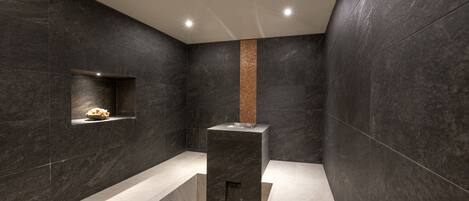 Un bagno moderno con pareti piastrellate in tonalità scura, una colonna centrale e un piccolo ripiano con un oggetto decorativo.