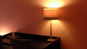 Ein schwach beleuchteter Raum mit Schreibtisch, Stuhl, Lampe und einem gerahmten Bild an der Wand.