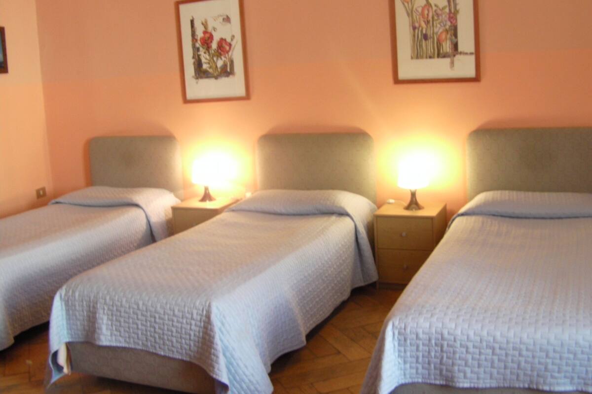 A hotel room with three single beds, each with a bedside table and lamp.