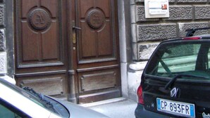 A double wooden door with a circular emblem, a stone wall with a sign, and a parked car. - Alla Stazione (Trieste)