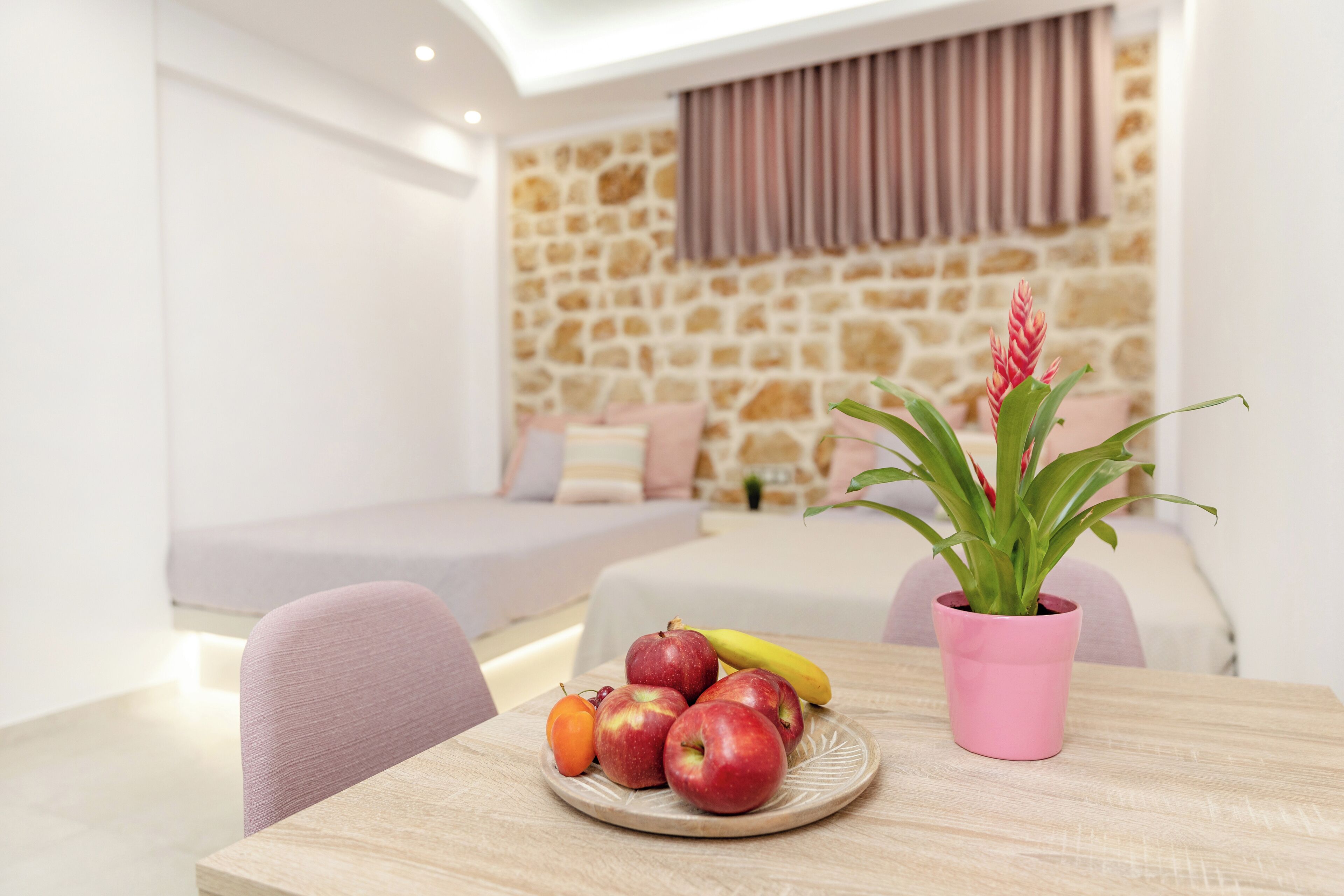 Uma mesa de jantar com uma travessa de fruta, uma planta em vaso e uma cama ao fundo.