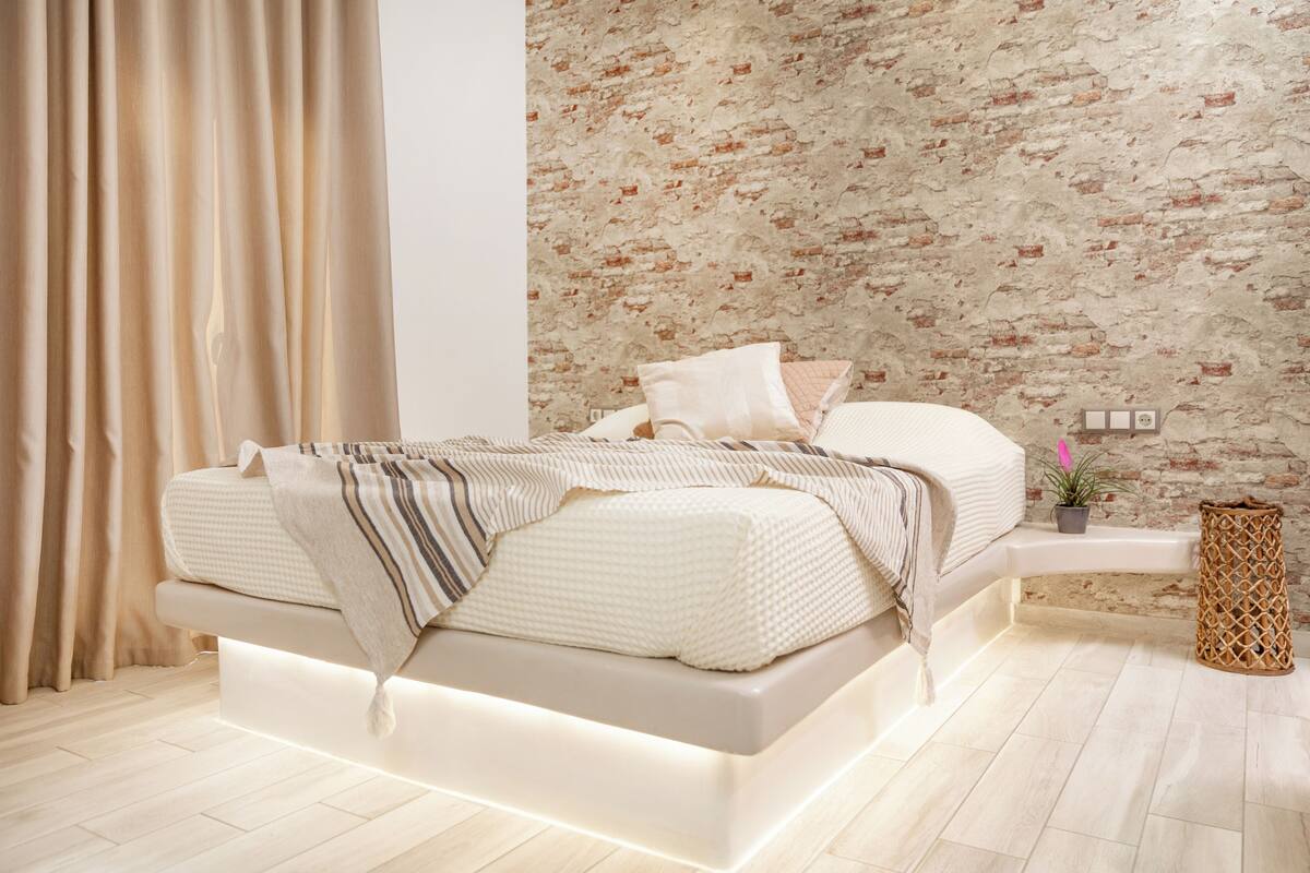 Un dormitorio moderno con cama, cortinas, pared de ladrillo, una planta pequeña y una cesta tejida.