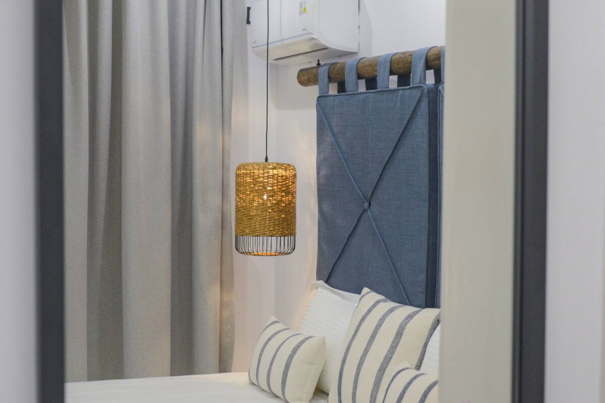 Una habitación de hotel moderna con una cama, una lámpara colgante, una ventana con cortinas y un espejo que refleja parte de la habitación.