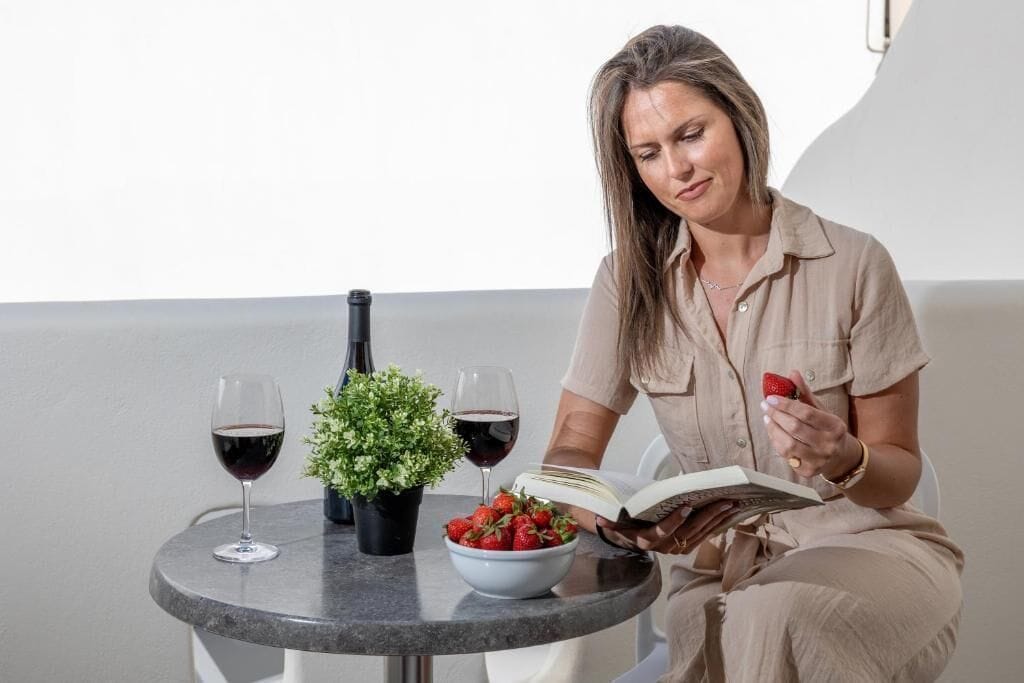 A person sitting at a round table with a book, a bowl of strawberries, two glasses of red wine, and a small potted plant.