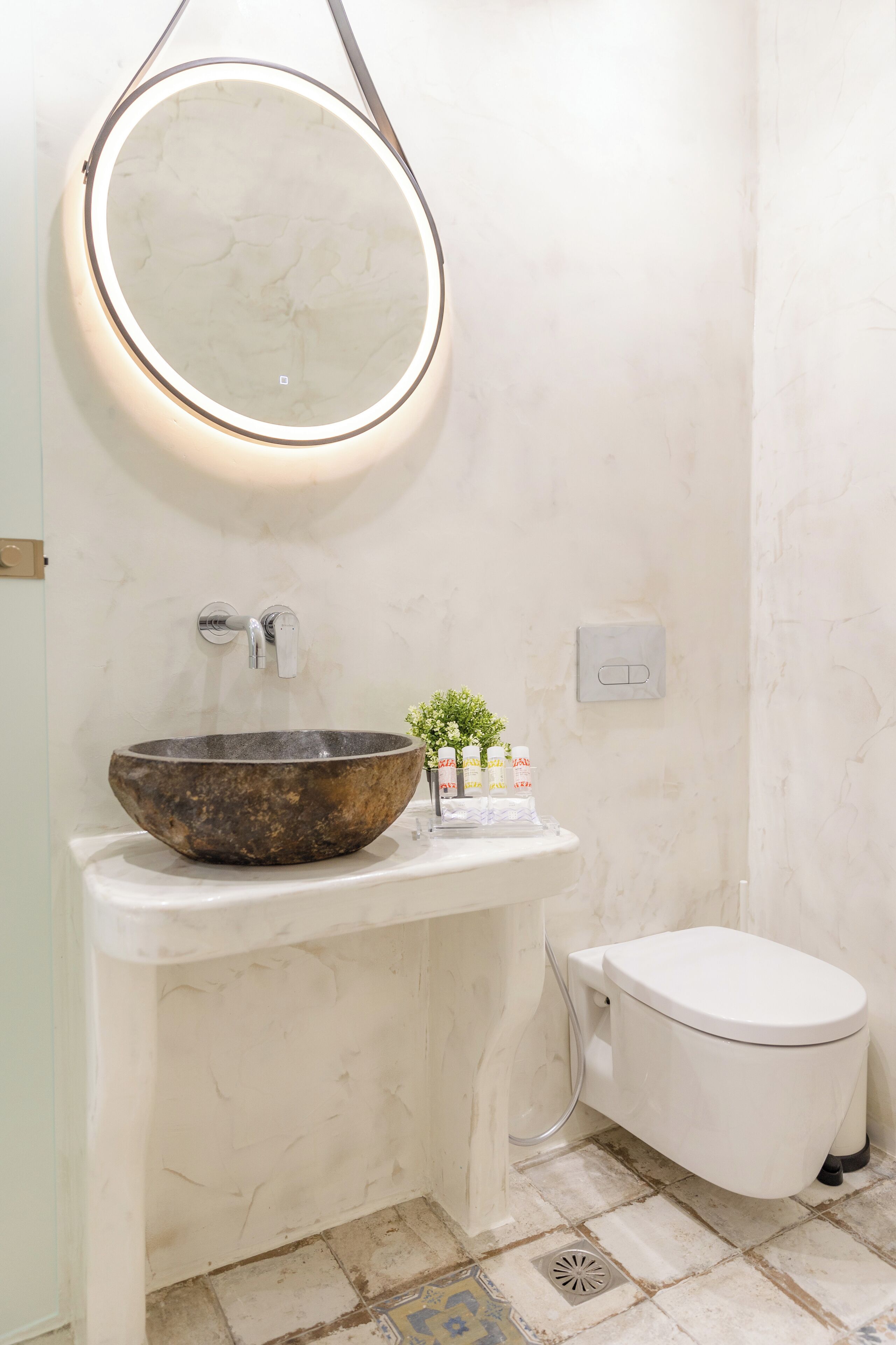 A bathroom with a marble sink, a round mirror, a toilet, and a small plant on the counter.