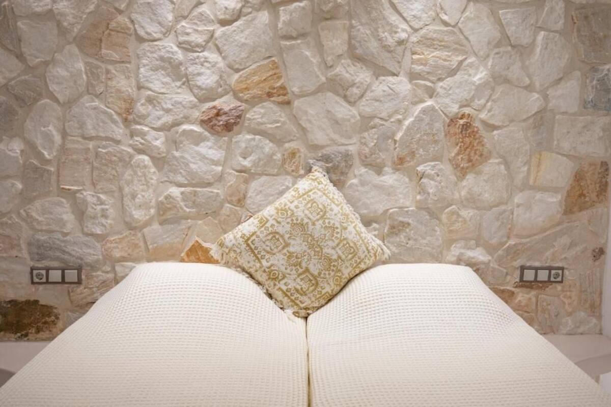 Una habitación con pared de piedra, una cama con ropa de cama blanca y una almohada estampada.