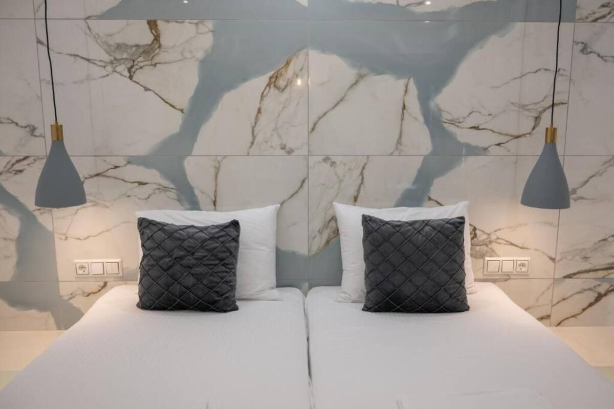 Una cama doble con dos almohadas acolchadas negras, una pared con estampado de mármol y dos lámparas colgantes.
