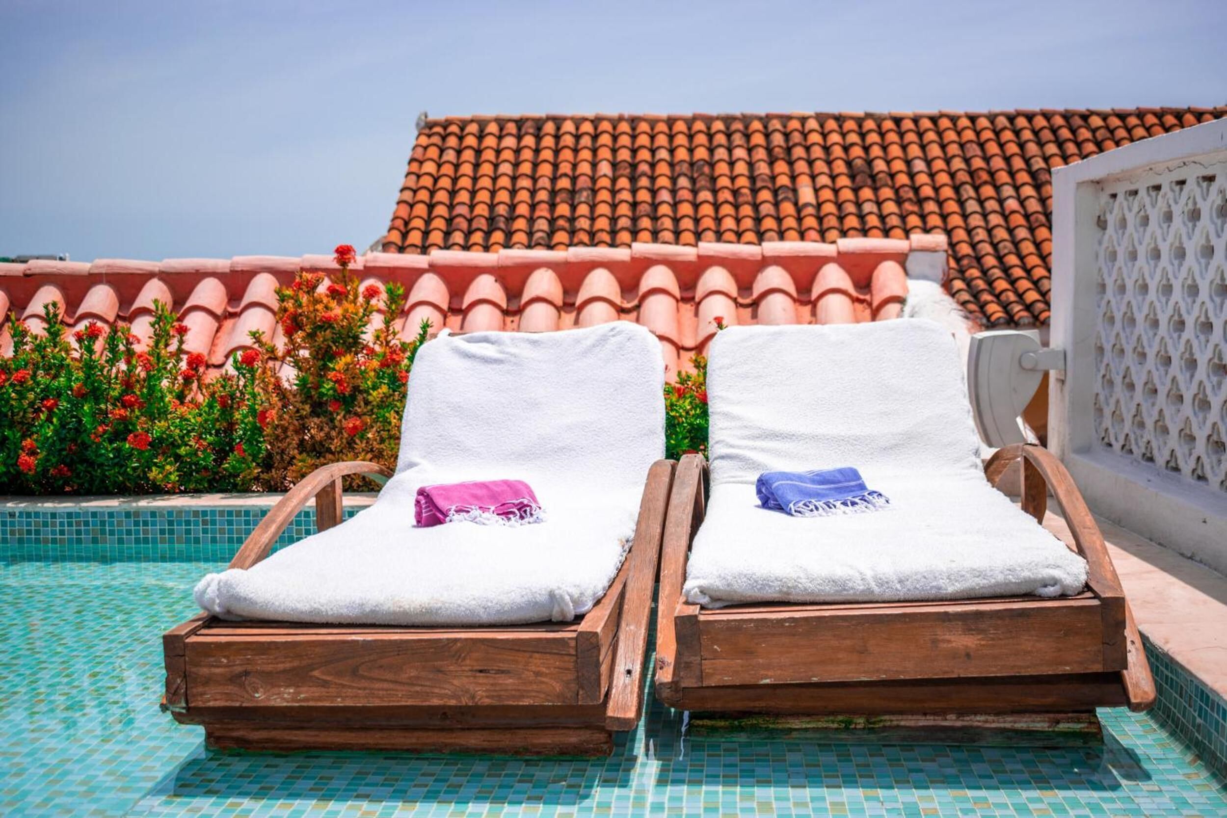 Two wooden lounge chairs with white towels and a pink and blue towel on them, positioned by a pool with a tiled floor and a tiled roof in the background.