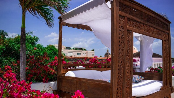 A wooden four-poster bed with a canopy, surrounded by vibrant pink flowers and a palm tree.