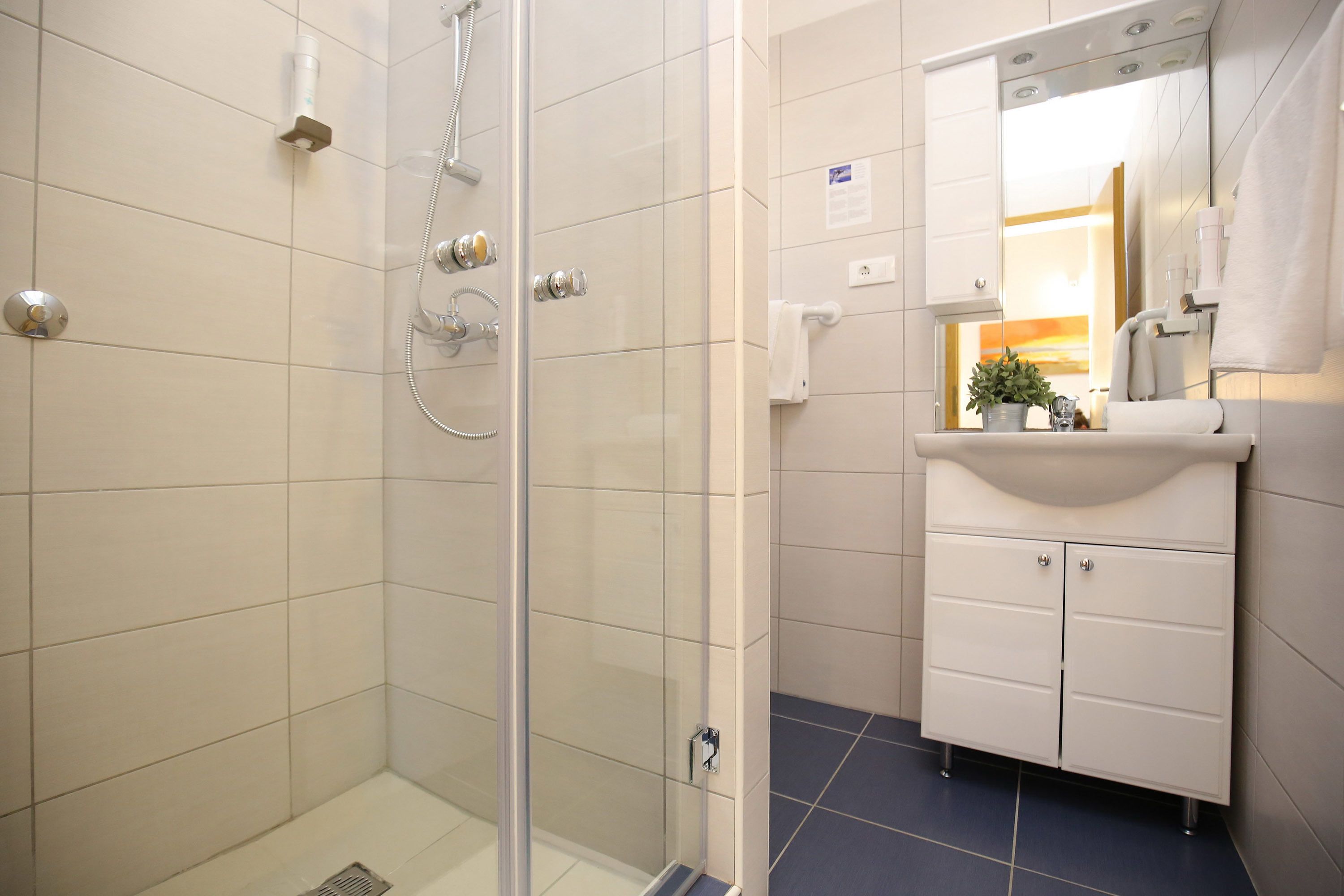 Un baño moderno con ducha envuelta en cristal, tocador blanco con espejo y suelo de baldosas azules.