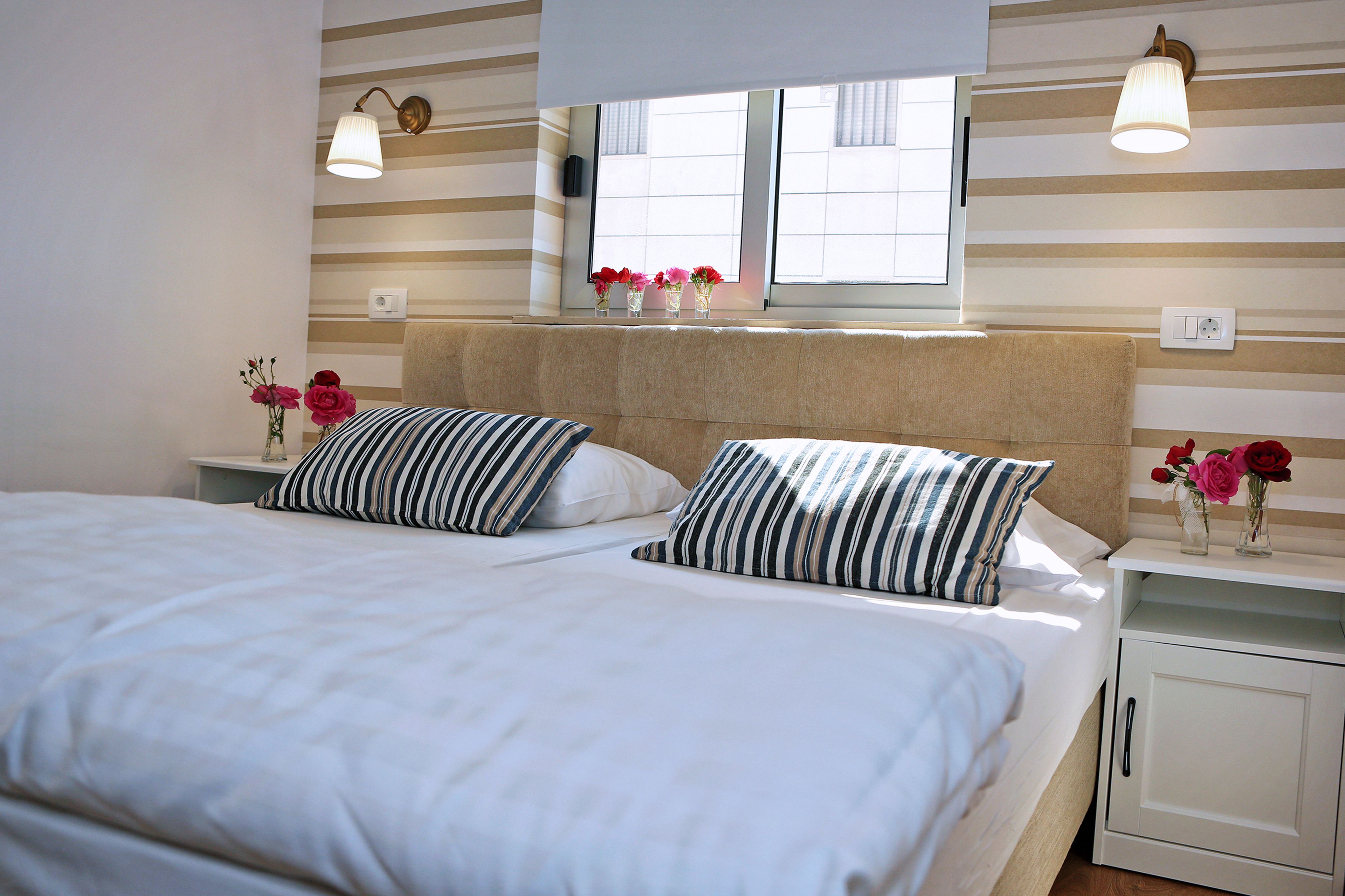 Una cama doble con almohadas a rayas, una mesita de noche con un florero y una ventana con vistas a edificios.