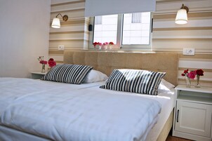 Una cama doble con almohadas a rayas, una mesita de noche con un florero y una ventana con vistas a edificios.