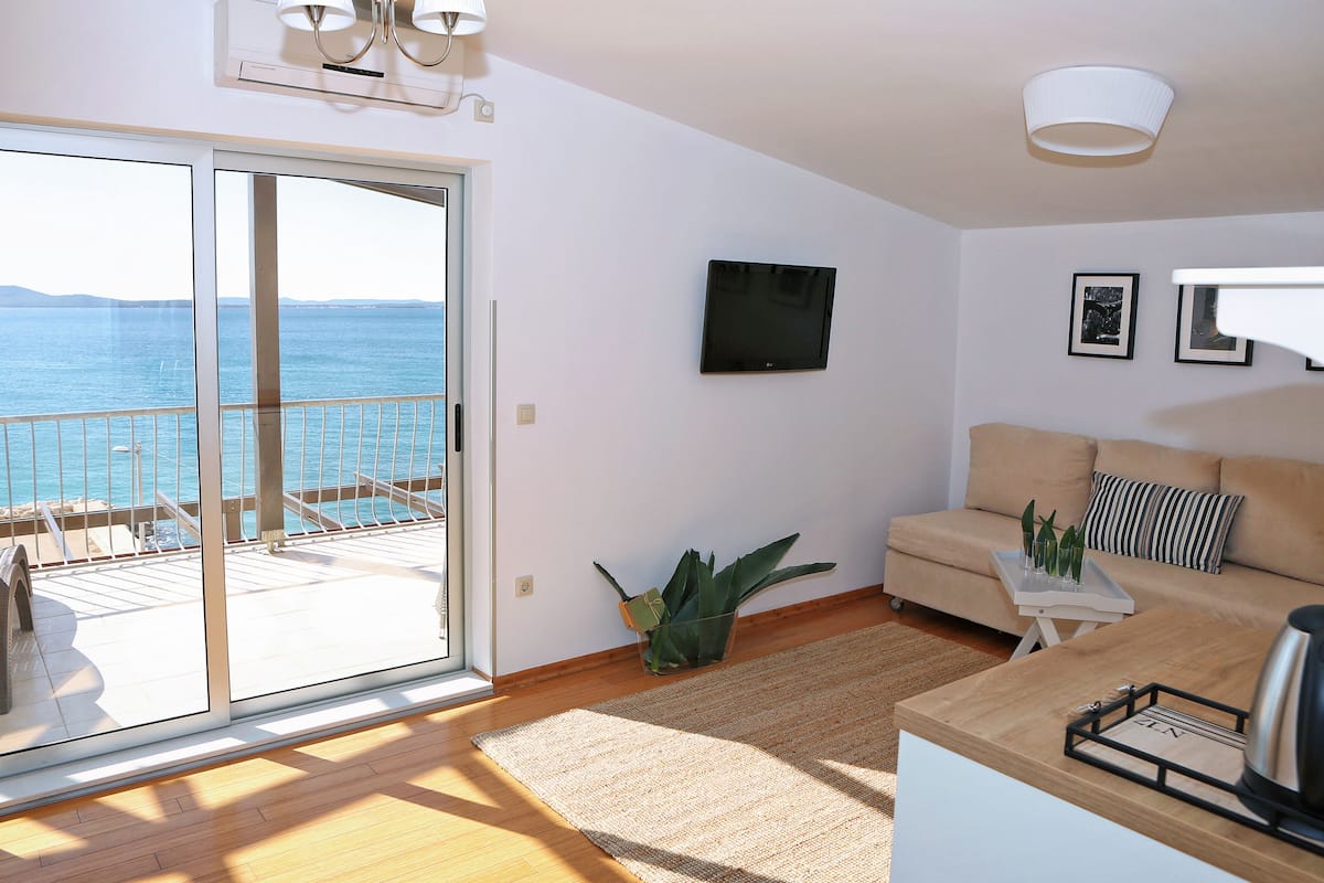 Ein modernes Wohnzimmer mit Sofa, Couchtisch und Fernseher. Ein Balkon bietet Blick auf das Meer.