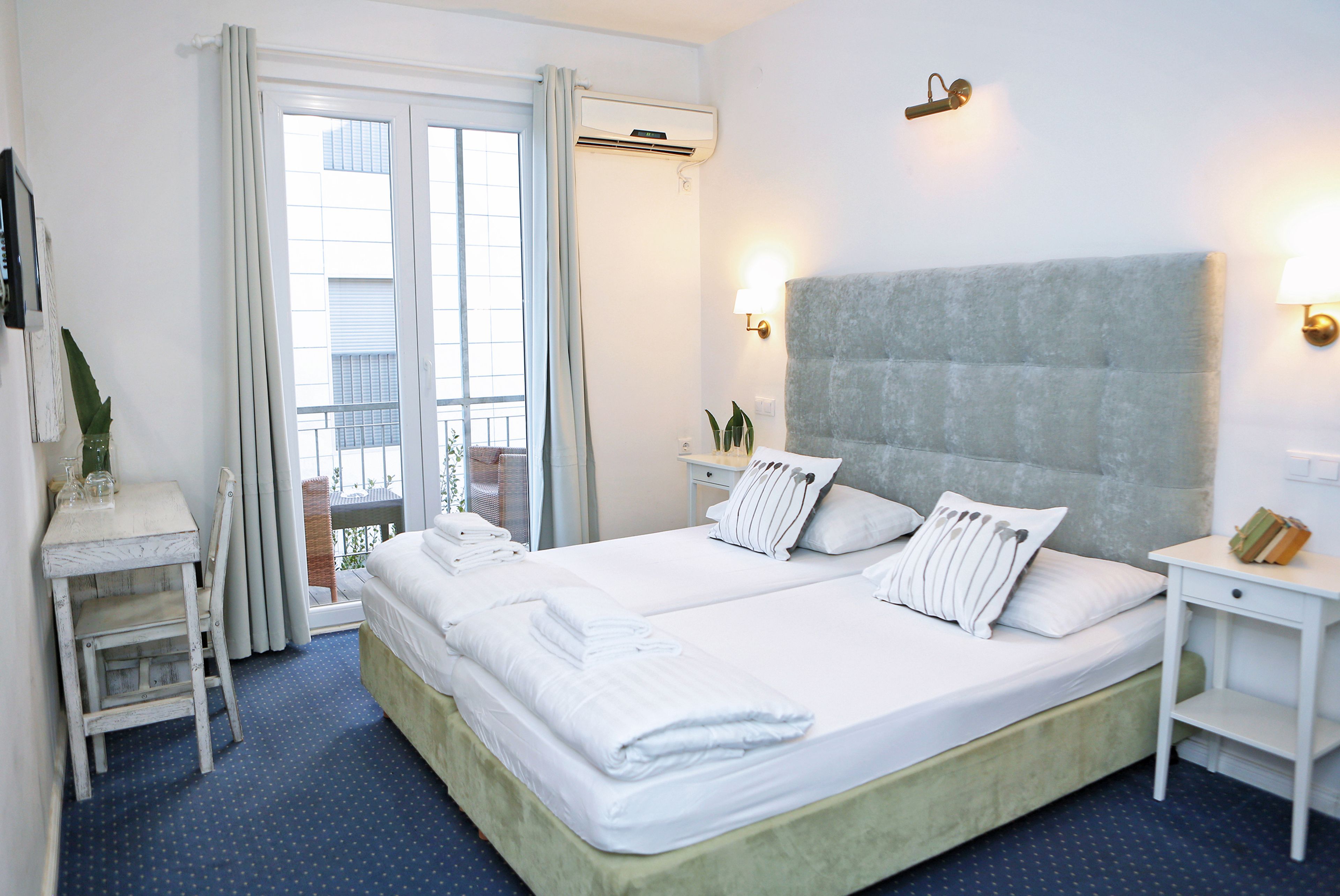 A double bed with white linens, a small bedside table, a nightstand, a chair, and a view of the outdoors through a window with curtains.