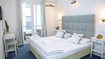 A double bed with white linens, a small bedside table, a nightstand, a chair, and a view of the outdoors through a window with curtains.
