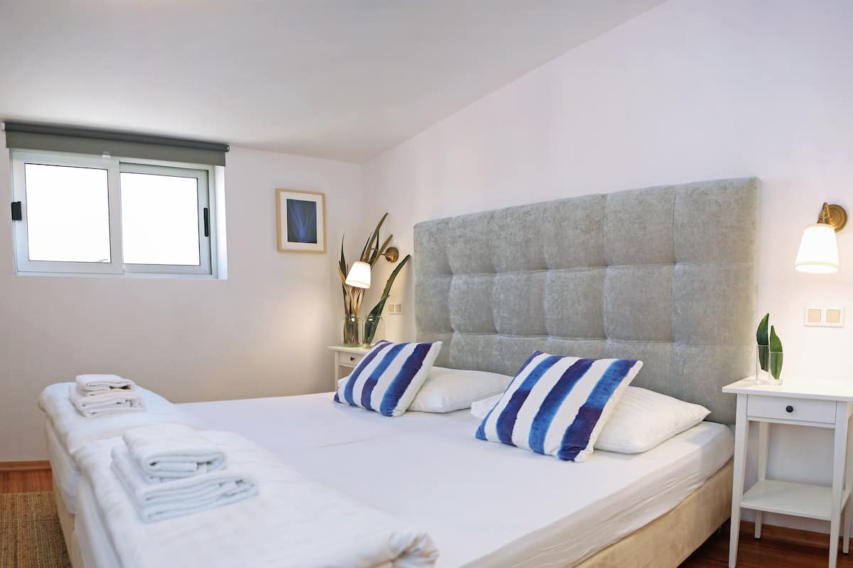 Ein ordentlich gemachtes Bett mit weißen Leinen und gestreiften Kissen, ein Nachttisch mit Lampe sowie ein Fenster mit Jalousien.