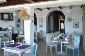 Un restaurante con sillas blancas y verdes, una mesa de comedor preparada con platos, vasos y cubiertos. Hay un televisor montado en la pared, así como varias fotos y cestas enmarcadas.