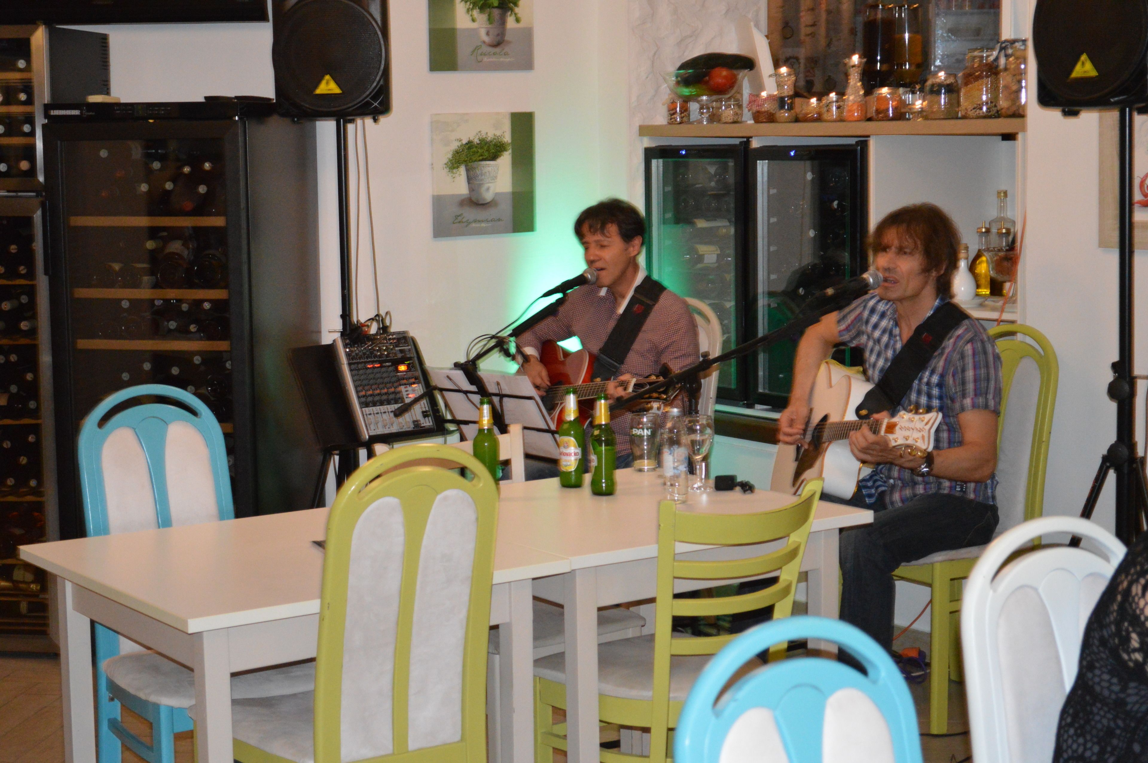 Een man speelt gitaar en zingt in een microfoon aan een tafel met een wijnfles en glazen.