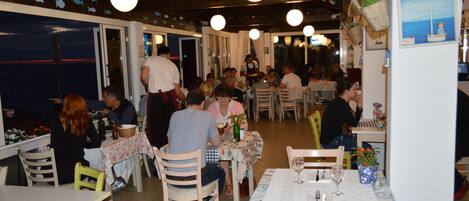 Un restaurante con varias mesas y sillas, vistas al mar a través de las ventanas y lámparas colgantes.