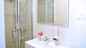 Une salle de bain avec une douche, un lavabo, un miroir et un petit vase avec une fleur.