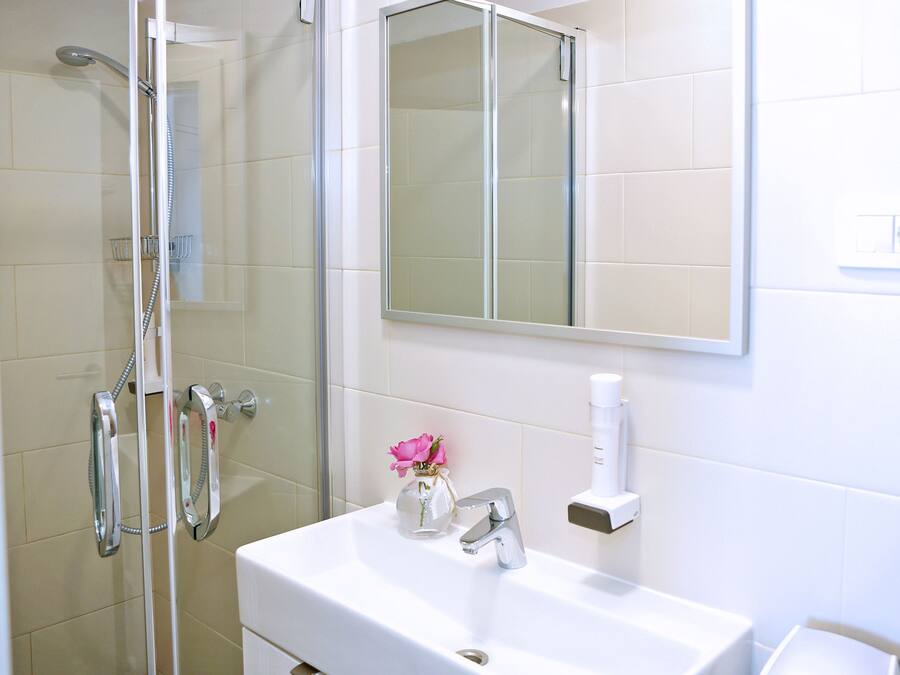 Un baño con ducha, lavamanos, espejo y un pequeño florero con una flor.