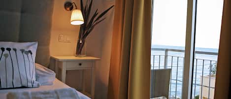 Una habitación de hotel con cama, mesita de noche, un florero con plantas y vista al mar desde un balcón.