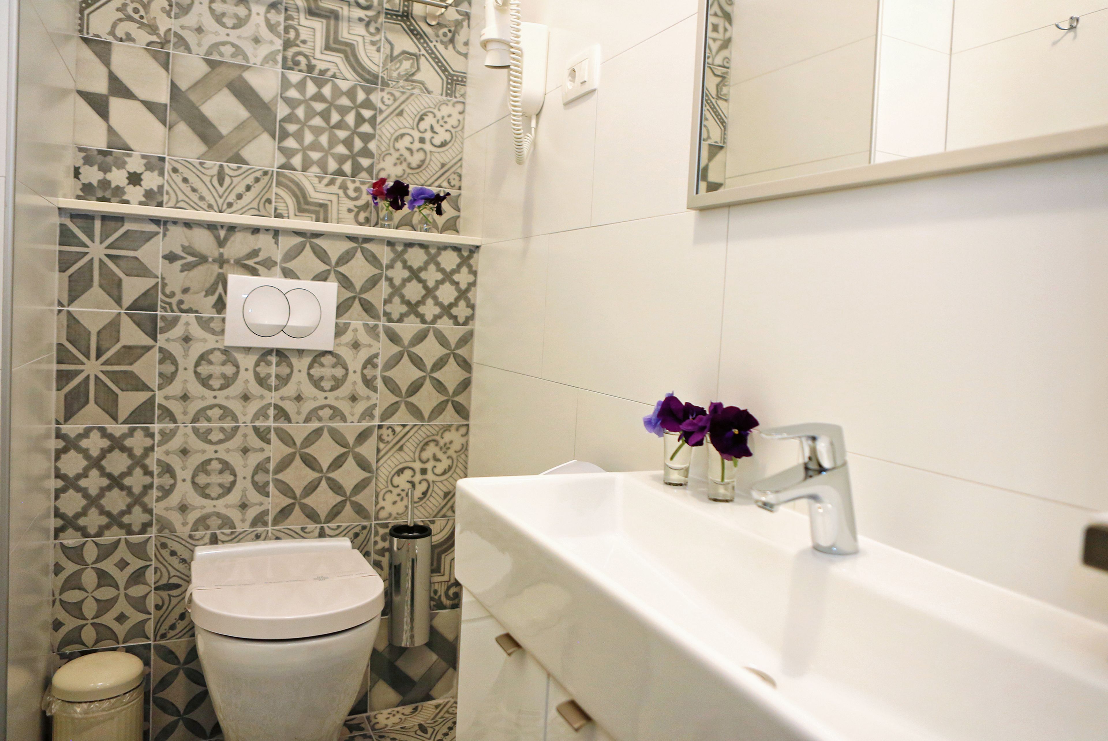 A bathroom with patterned tiles, a toilet, a bidet, a sink with a mirror, and two vases with flowers.