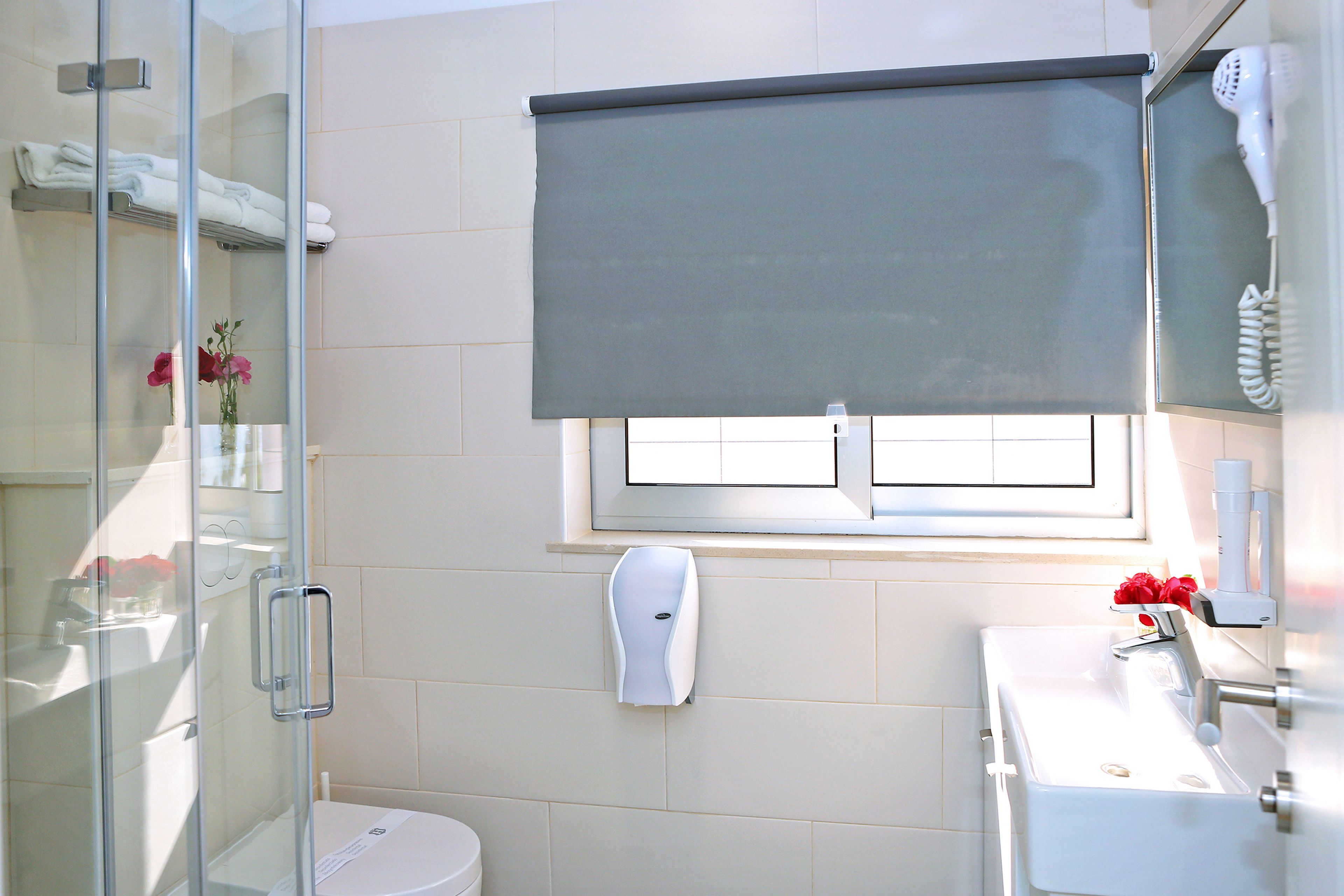 Une salle de bain avec une douche à parois de verre, un lavabo blanc, un distributeur de serviettes et une fenêtre avec un store.