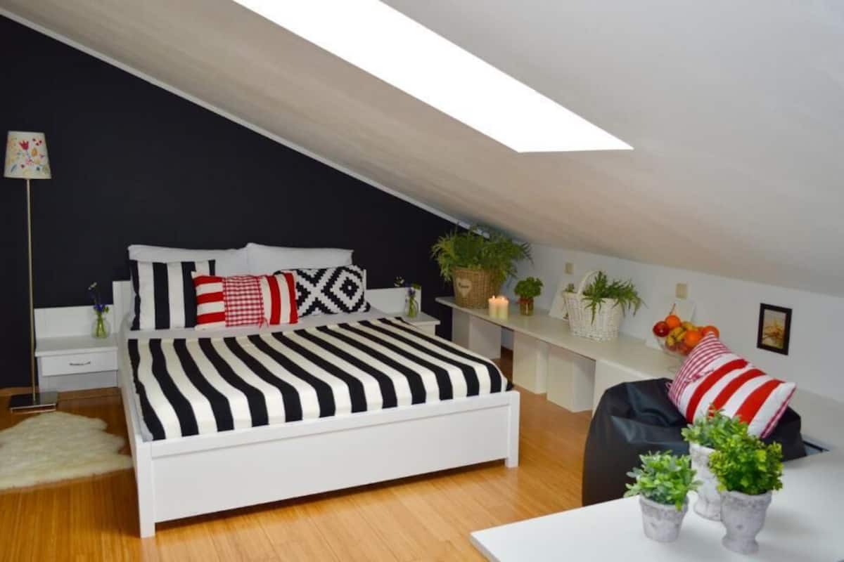 Ein modernes Schlafzimmer mit schrägen Wänden, einem weißen Bett mit gestreifter Bettwäsche, einem Nachttisch, einer Lampe und einer kleinen Pflanze.