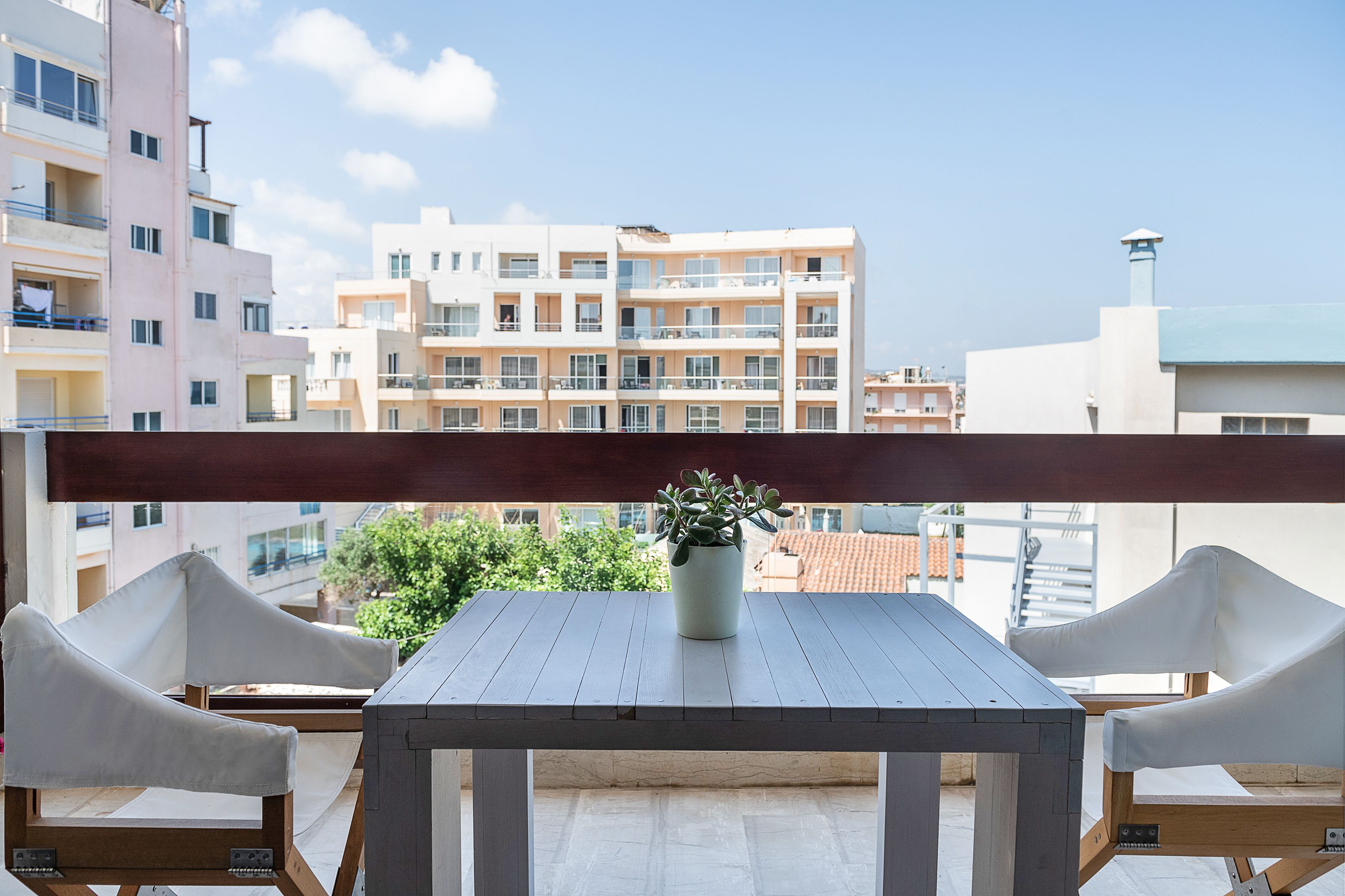 Un balcón con una mesa de madera y dos sillas blancas, una planta en maceta y vistas a edificios de varios pisos.