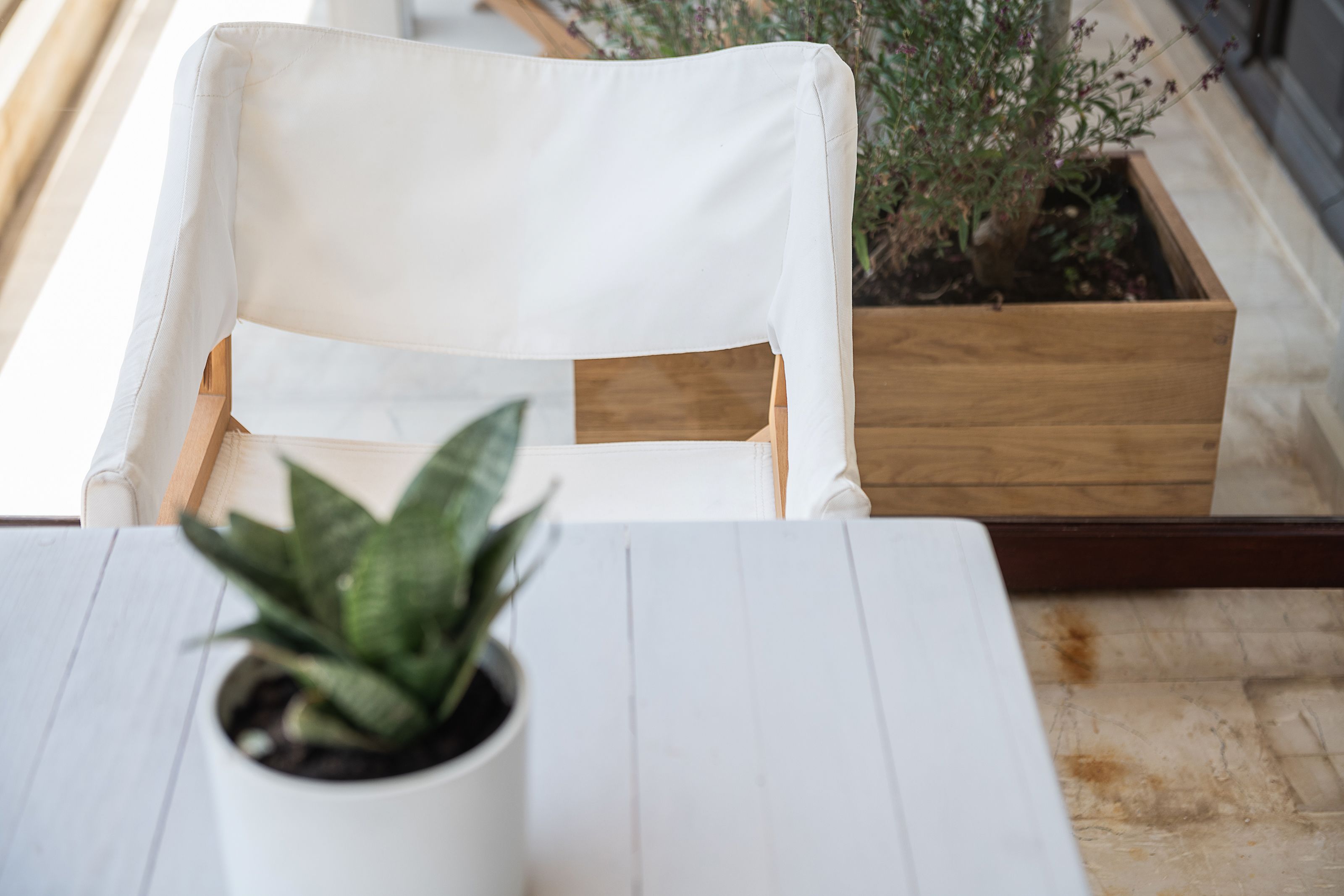 A white cushioned chair with a wooden frame, a potted plant on a table, and a wooden planter with a plant.