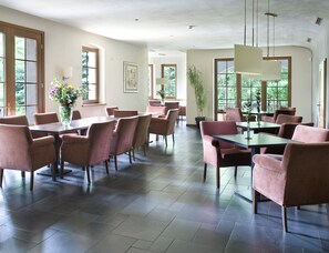 Een ruime eetruimte met meerdere tafels en stoelen, grote ramen die natuurlijk licht binnenlaten, en een modern plafondontwerp.