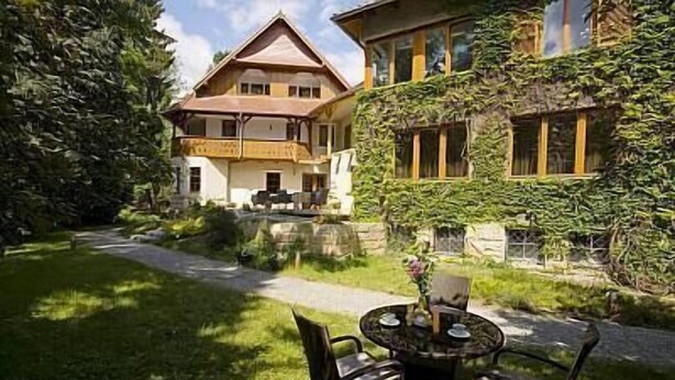 Ein zweistöckiges Haus mit Balkon, umgeben von üppiger Vegetation und einem gepflasterten Weg, der zu einem runden Tisch mit Stühlen führt.