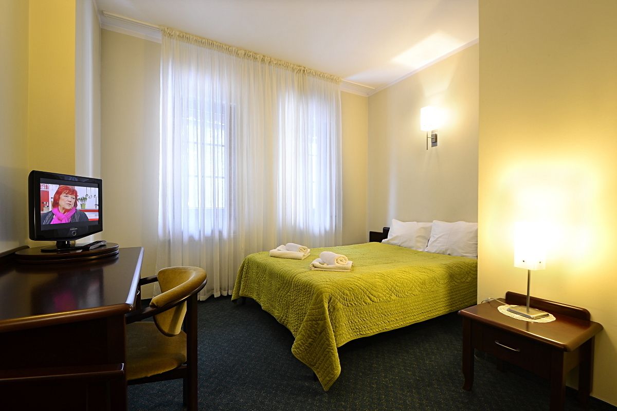 A hotel room with a bed, a desk with a computer, a chair, a bedside table with a lamp, and a window with curtains.