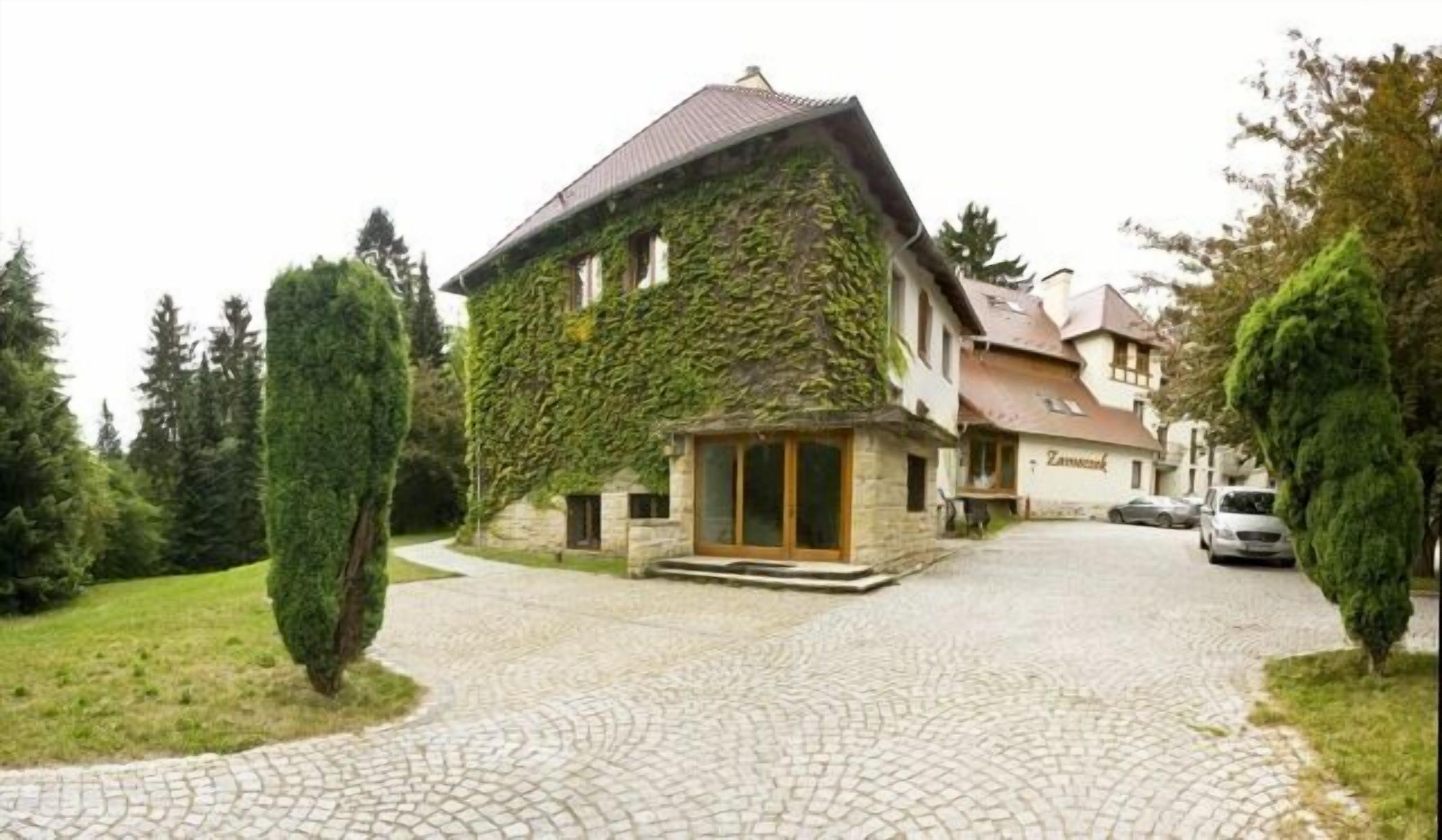 A house with a stone facade and ivy-covered walls, a paved driveway, and a car parked in the background.