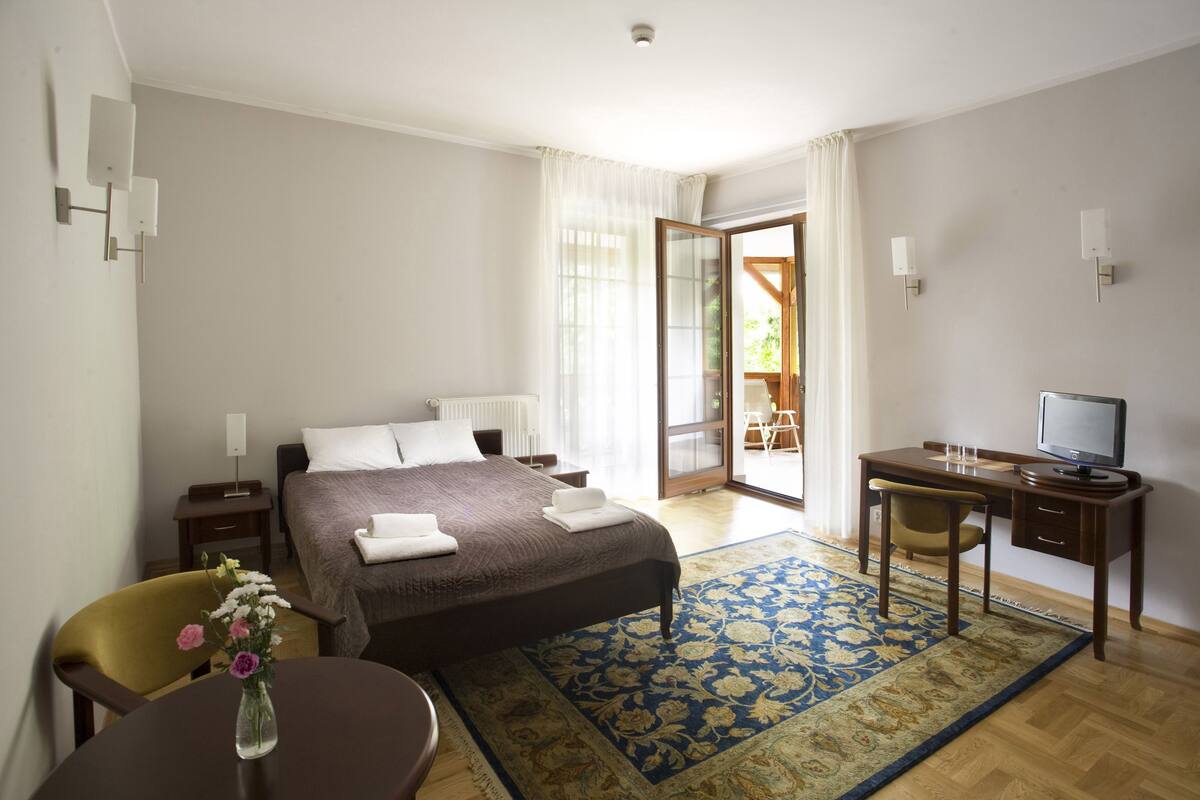 Habitación de hotel con cama, escritorio, silla, pequeña mesa con un florero y vistas al exterior.