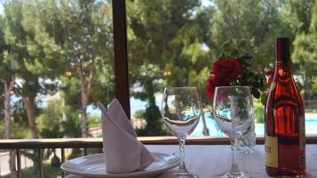 Una mesa dispuesta con un plato, una copa de vino y una botella, con vistas a árboles y una piscina al fondo.