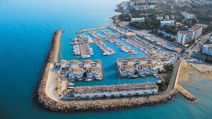 An aerial view of a coastal marina with multiple docks and boats, surrounded by residential buildings and a rocky breakwater.