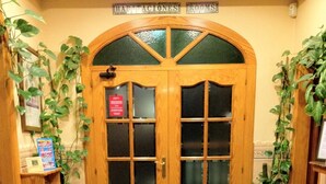 Entrada de hotel con puerta doble de madera, alfombra con símbolo 'HC' y plantas en macetas a ambos lados.