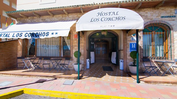 Entrada de un hotel con un cartel que dice "Hostal Los Corchos" y zona de estar al aire libre con mesas y sillas.