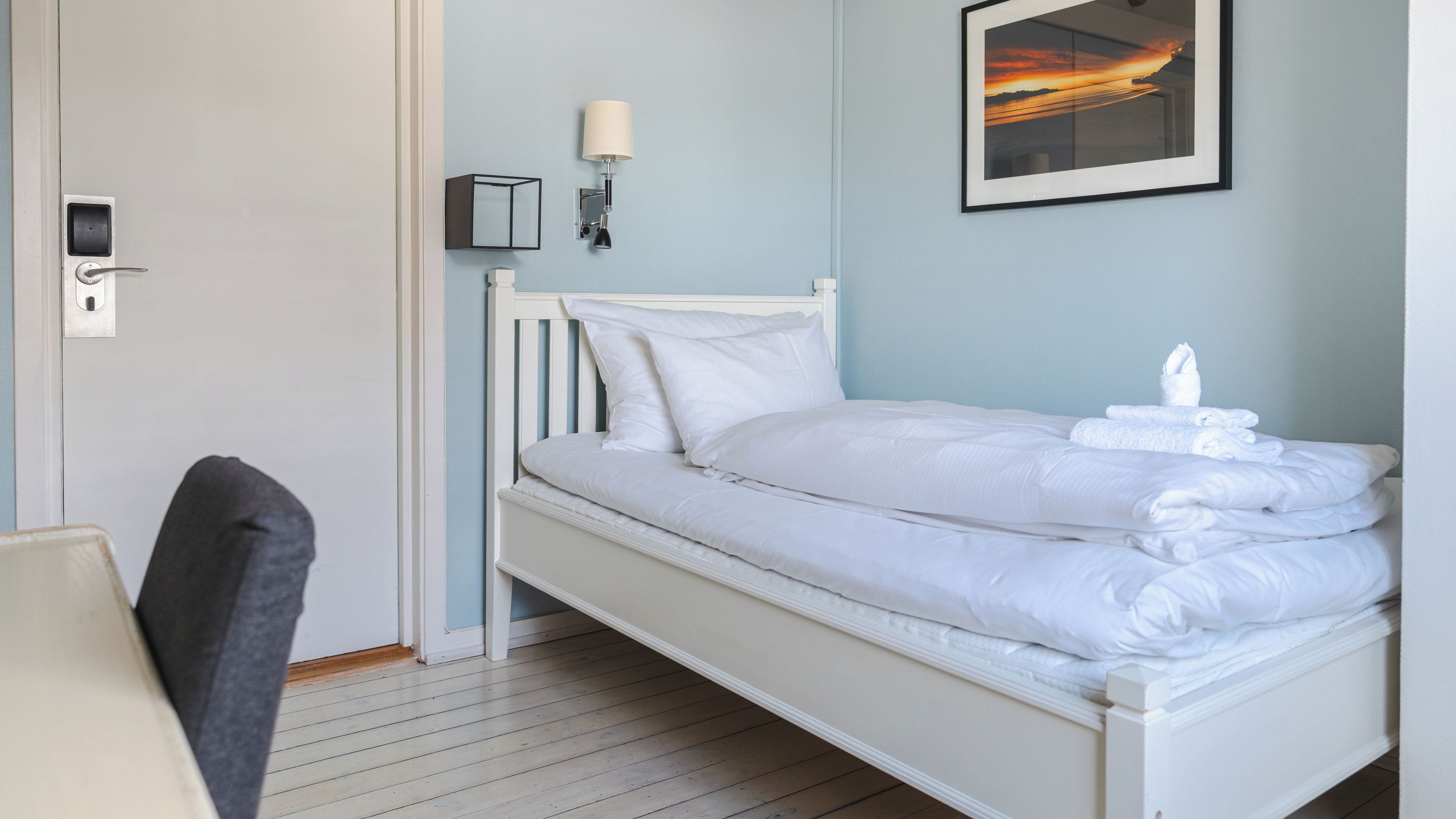 A single-bed room with a white headboard, a bedside table, a wall-mounted lamp, and a framed picture above the bed.