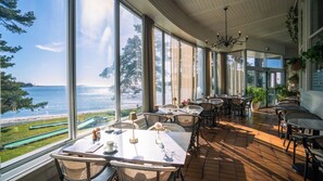 Un comedor soleado con mesas y sillas, ventanas amplias con vista al mar y un área con césped.