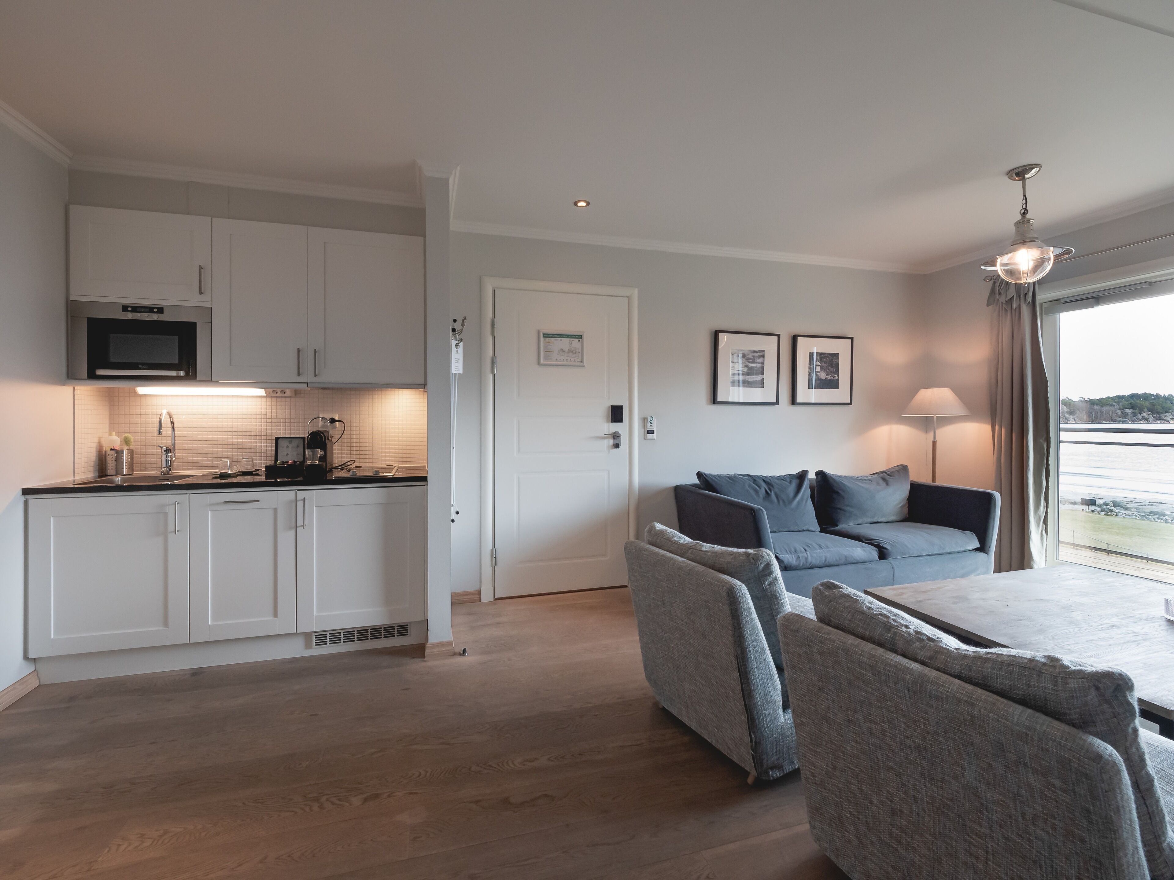 A modern kitchen with white cabinets, a microwave, and a coffee maker. There is a grey sofa with cushions in a living area, and a dining table with chairs. A large window offers a view of the water.