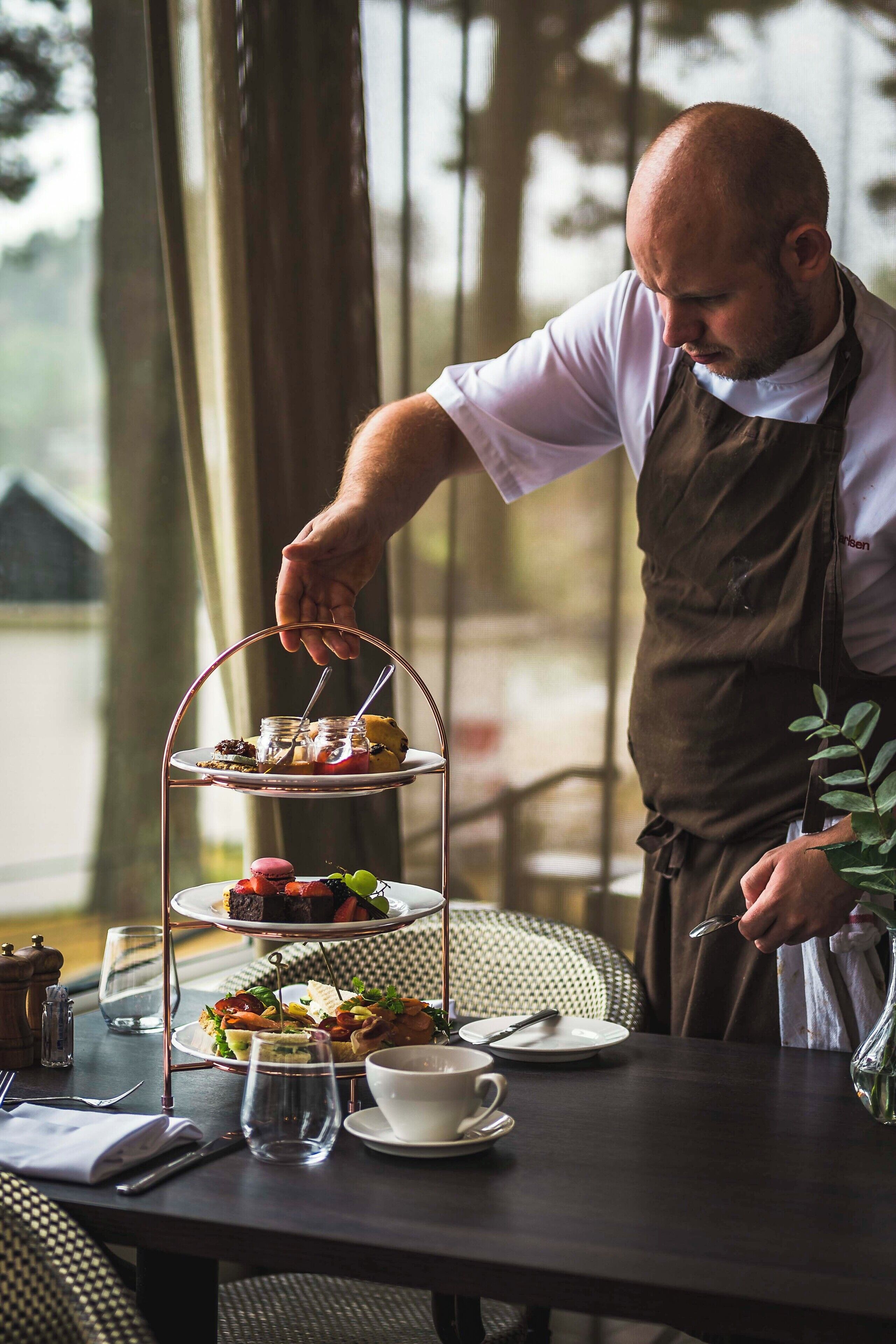 A person serving a three-tiered dessert tray on a table with a cup and saucer, salt and pepper shakers, and a glass.