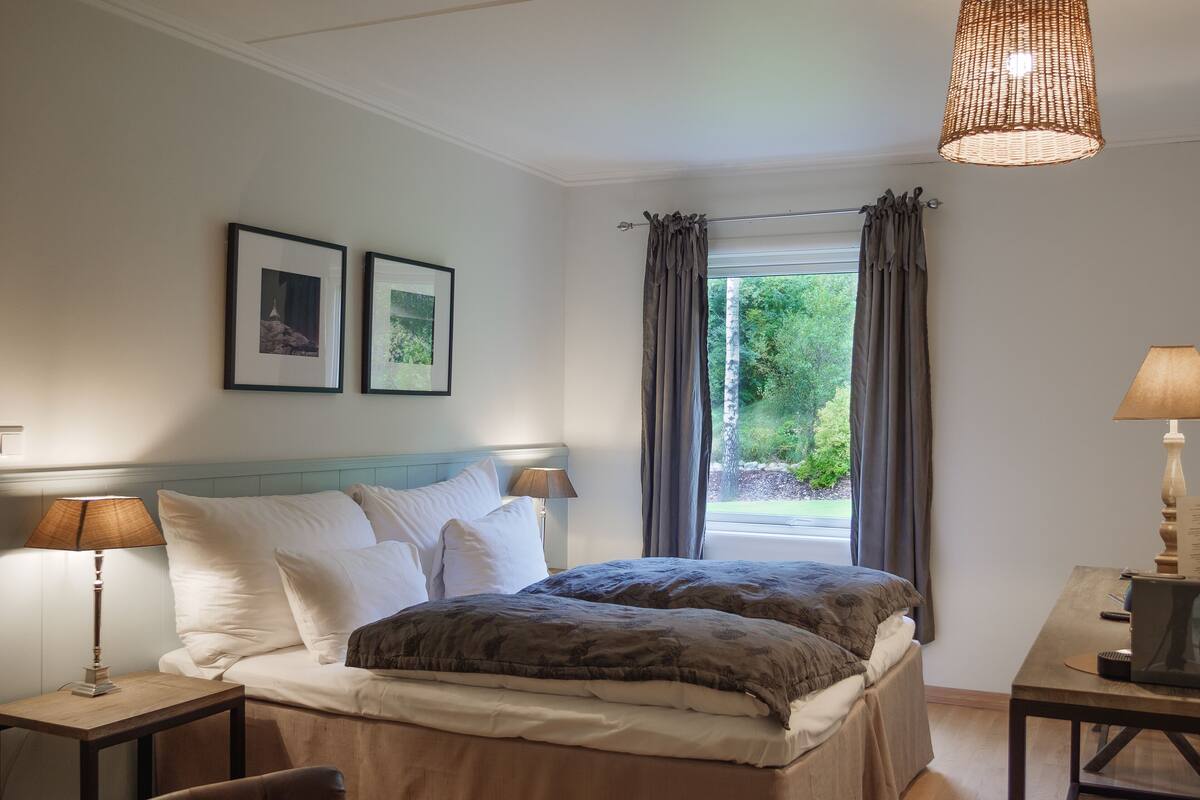 Un dormitorio con cama, lámparas de noche, una silla, una mesa y una ventana con cortinas.