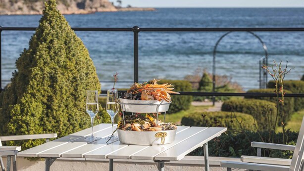 Un plat de marisc sobre una taula, amb un got de vi blanc, amb vista al mar i un arbre de fulles perennes i una barana metàl·lica.